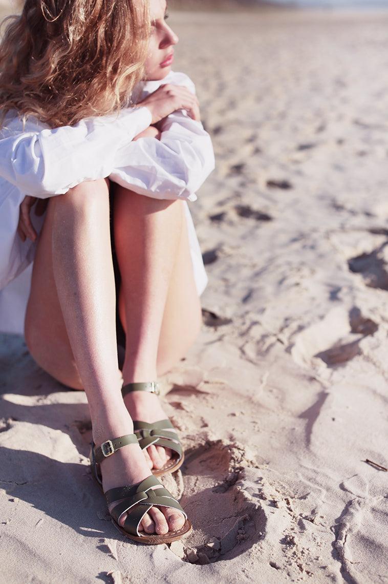 An adult with long blonde hair is sitting on the beach wearing a white cover up with their arms crossed over their knees.  They are wearing our Olive Salt Water Original .